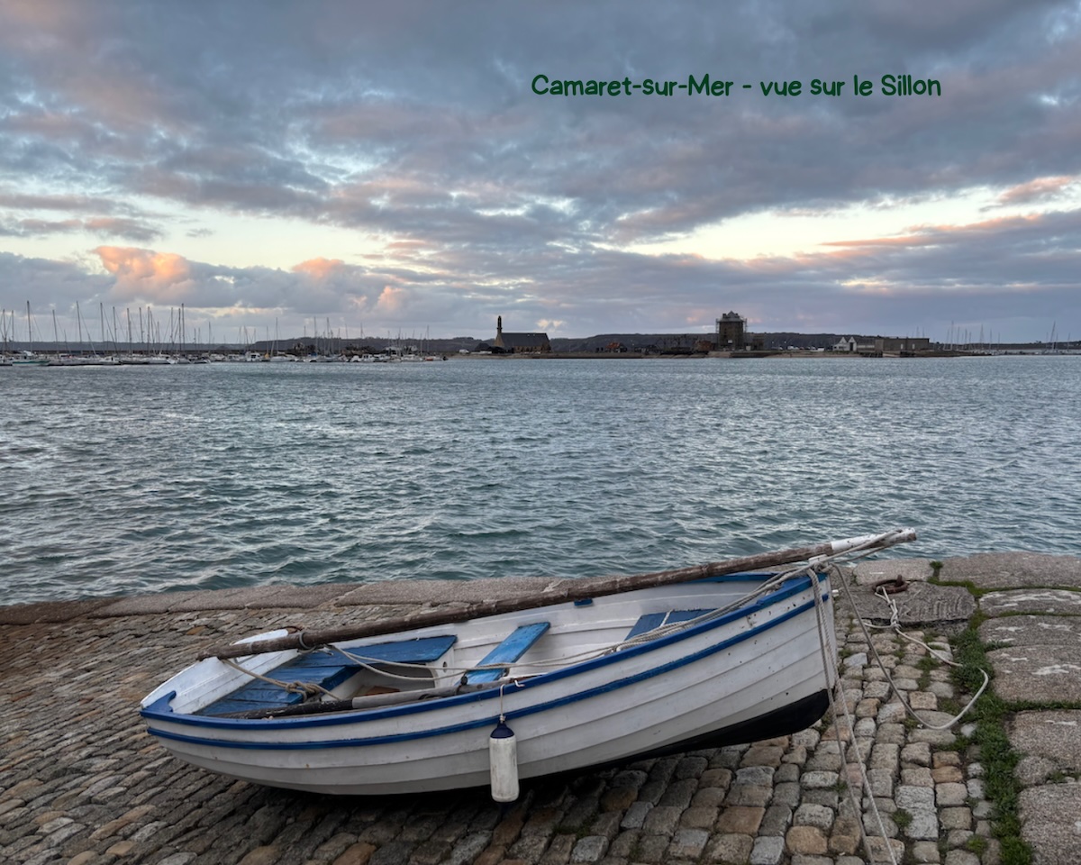 Barque blanche et bleue échouée sur une cale pavée à Camaret-sur-Mer au crépuscule, avec le port et le sillon en arrière-plan.