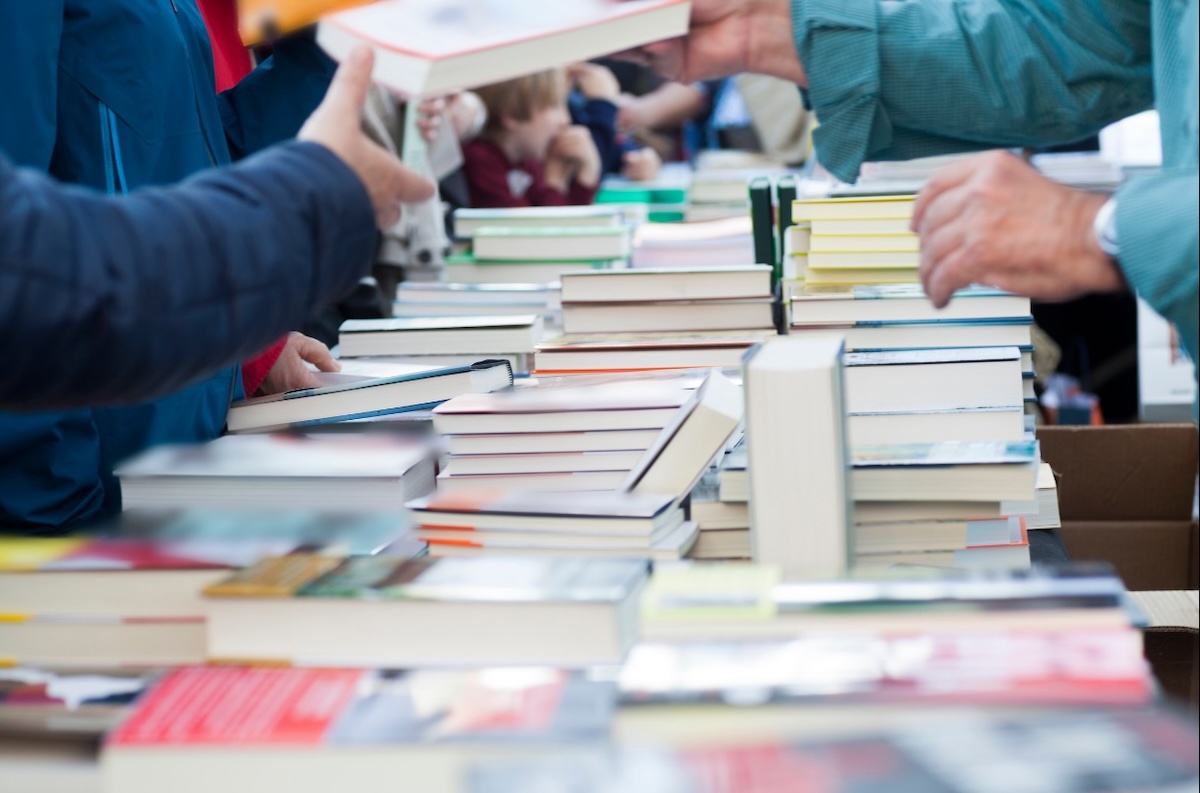 Livres empilés sur une table lors d’un salon du livre, avec des visiteurs feuilletant les ouvrages